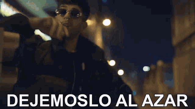 a man wearing sunglasses is standing in front of a building with the words dejemoslo al azar written below him .