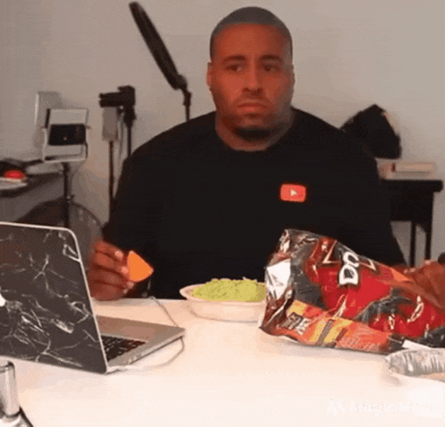 a man is sitting at a table with a laptop and a bag of doritos .