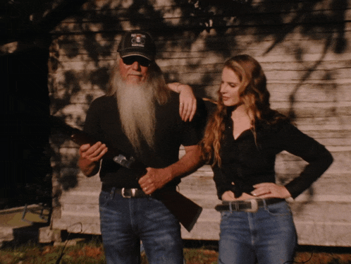 a man with a beard holds a gun next to a woman with long hair