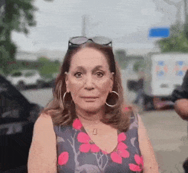 an older woman wearing a floral dress and hoop earrings is standing in front of a car .