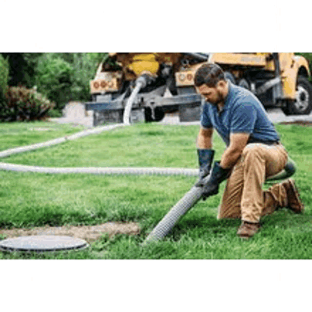 a man is kneeling down in the grass while a septic tank is being pumped .