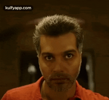 a man with a beard and a red shirt is looking at the camera with a serious look on his face .