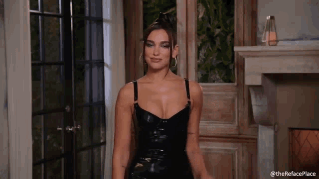 a woman in a black latex dress is standing in front of a fireplace in a room .