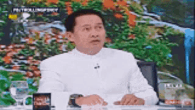 a man in a white shirt is sitting at a table talking on a television show .