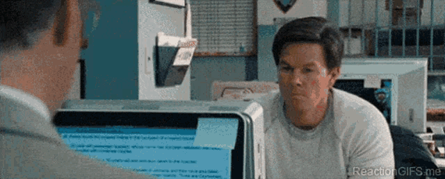 a man is sitting at a desk in front of a computer and looking at another man .