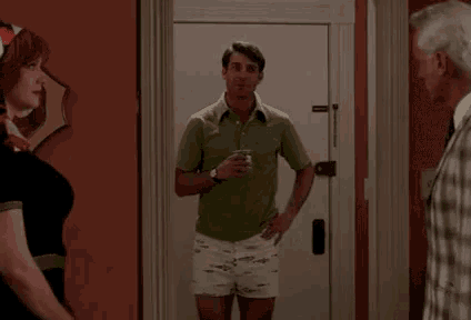 a man in a green shirt and white shorts is standing in front of a door holding a cup of coffee .