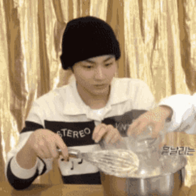 a man wearing a black beanie is holding a whisk over a bowl of food .