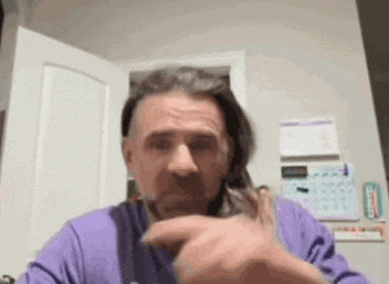 a man in a purple shirt is cutting his hair with a pair of scissors while sitting at a table .