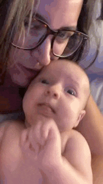 a woman wearing glasses is holding a baby in her arms and kissing it on the forehead .