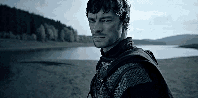 a man in armor stands on a beach near a lake