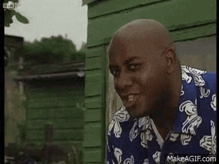 a bald man in a blue and white shirt is standing in front of a green shed .