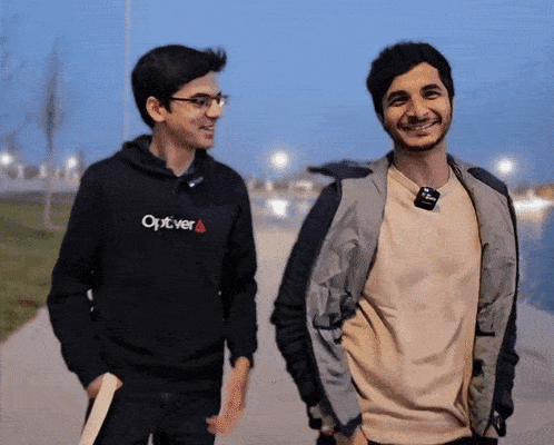a man wearing a black optiver sweatshirt smiles while walking with another man