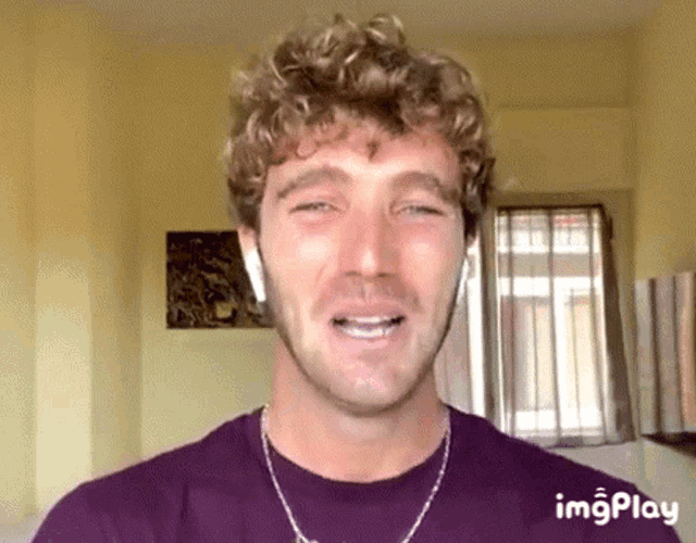 a man with curly hair is wearing a purple shirt and a necklace and smiling .