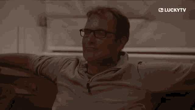 a man wearing glasses is sitting on a couch with his arms outstretched and a lucky tv logo in the background .
