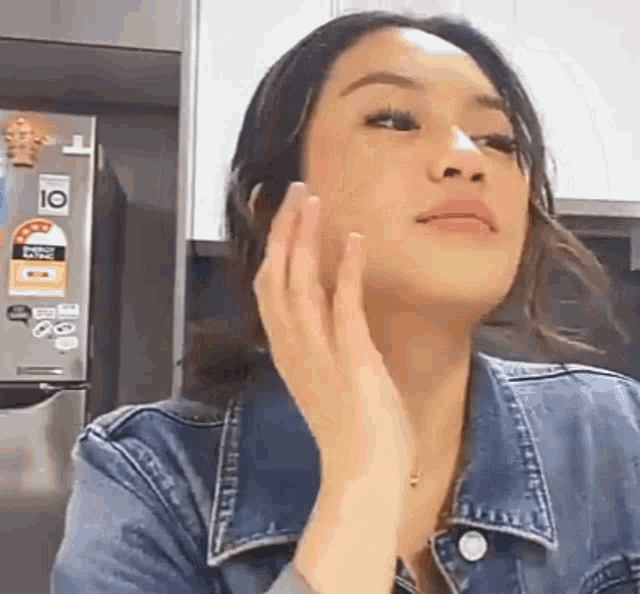 a woman in a denim jacket is touching her face in front of a fridge .
