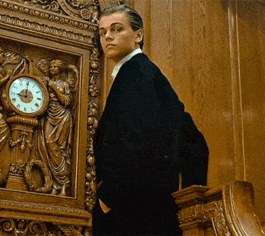 a man in a suit stands in front of a clock with roman numerals on the face