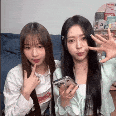 two girls are standing next to each other and one has a phone in her hand and the other has her finger on her lips