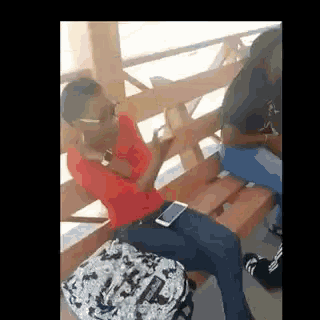 a man in a red shirt is sitting on a wooden bench with a backpack and a cell phone .