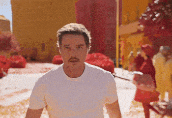 a man with a mustache wearing a white t-shirt stands in front of a crowd of people