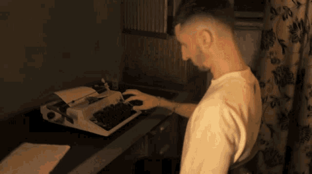 a man is typing on a typewriter with a floral curtain behind him
