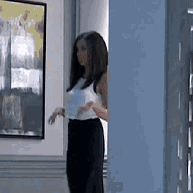 a woman in a white tank top and black pants is standing in front of a wall with a painting on it .