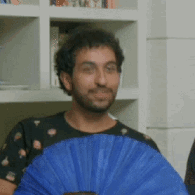 a man with a beard holds a blue fan in front of a bookshelf