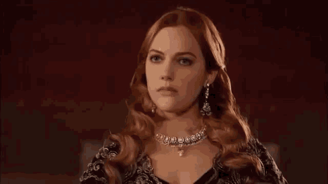 a woman with red hair is wearing a necklace and earrings and looking at the camera .