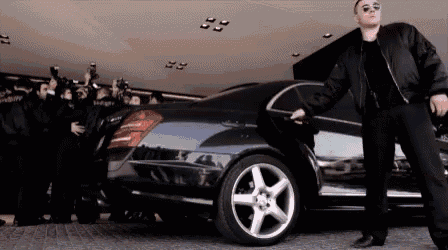 a man stands in front of a black mercedes with a crowd of people behind him
