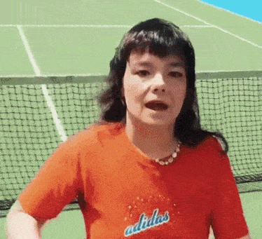 a woman wearing a red adidas shirt stands on a tennis court