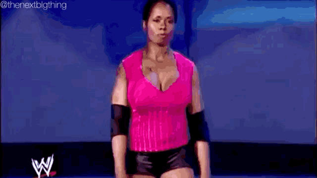 a woman in a pink top and black shorts is standing on a stage in front of a wrestling ring .