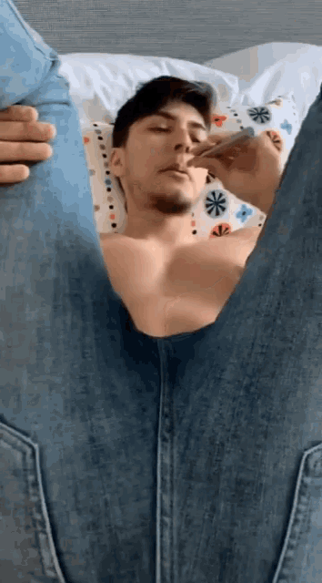 a shirtless man is laying on a bed with his legs crossed and smoking a cigarette
