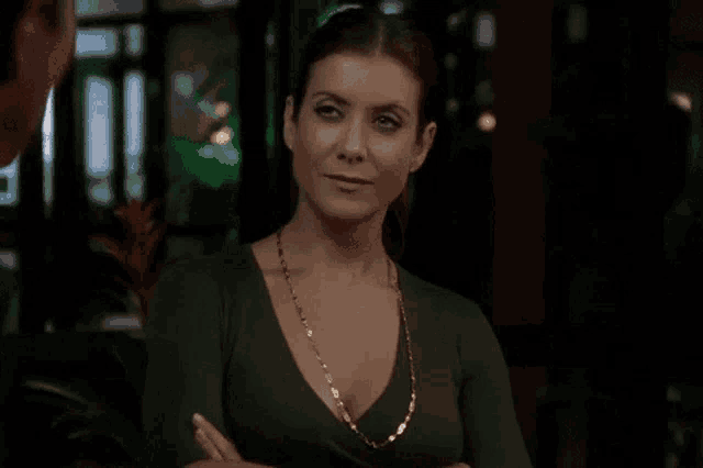 a woman wearing a green top and a gold necklace stands with her arms crossed