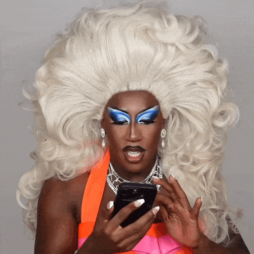 a drag queen with a large white wig is looking at her cell phone