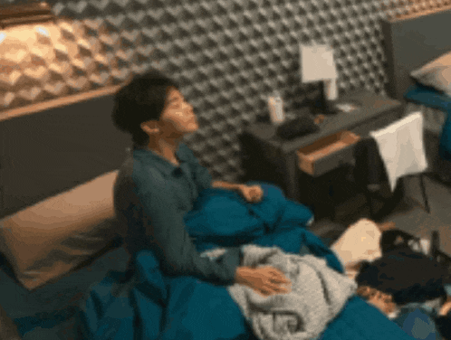 a man is sitting on a bed with a blue blanket while another man sleeps on the floor .
