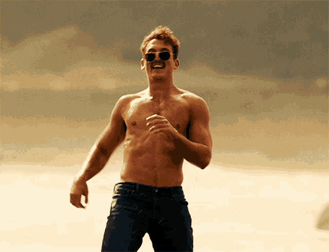 a shirtless man wearing sunglasses and jeans stands in the desert