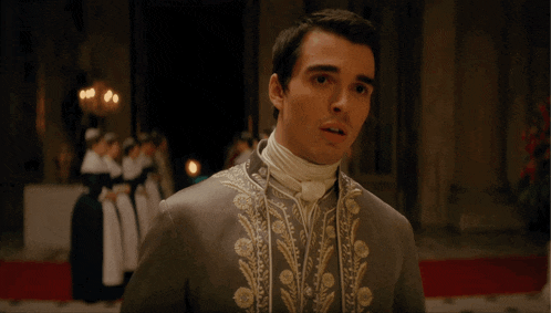 a man in a grey and gold embroidered coat stands in a dark room