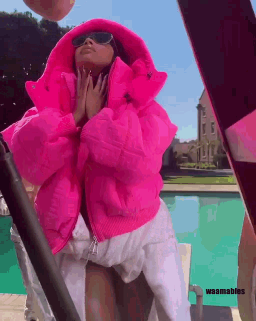 a woman wearing a bright pink jacket and sunglasses stands next to a swimming pool