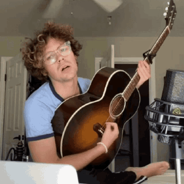 a man with glasses is holding a guitar in front of a microphone