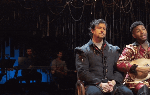 two men are sitting on a stage with one playing a lute