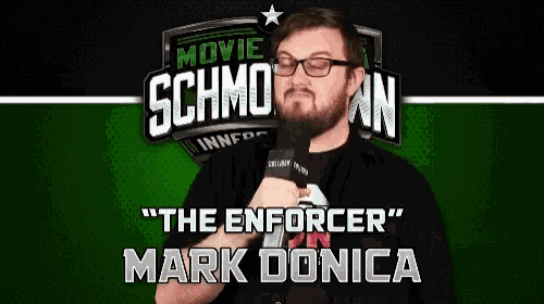 a man with glasses is holding a microphone in front of a sign that says " the enforcer mark donica "