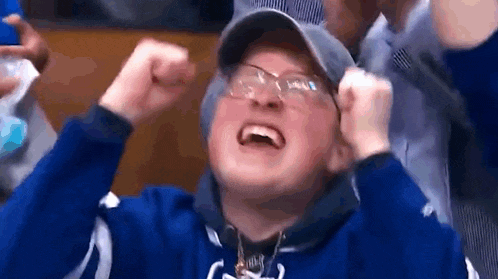 a man wearing glasses and a hat is sitting in a crowd with his hands in the air .