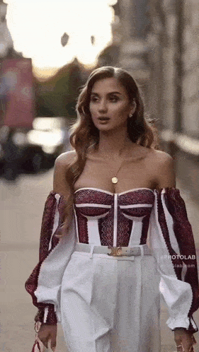 a woman is walking down a street wearing a burgundy and white off the shoulder top and white pants .