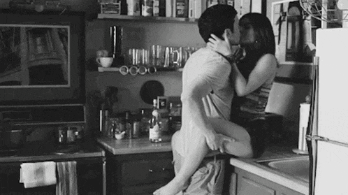 a man and a woman are kissing in a kitchen in a black and white photo .