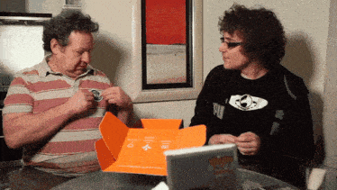 two men are sitting at a table looking at a box that says pop