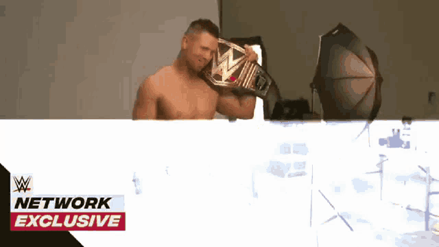 a shirtless wrestler is holding a wrestling belt in front of a network exclusive banner