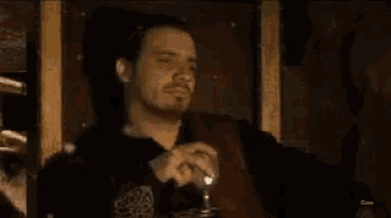 a man with a beard is sitting in a dark room holding a glass of wine .