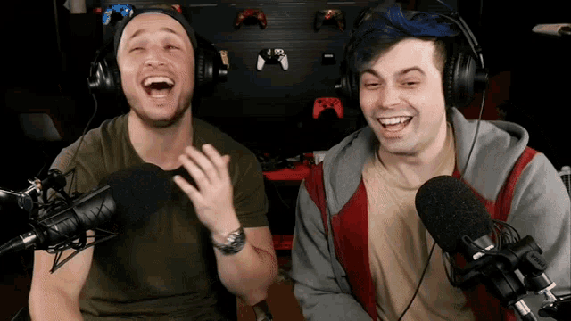 two men wearing headphones are laughing in front of microphones with xbox controllers on the wall behind them