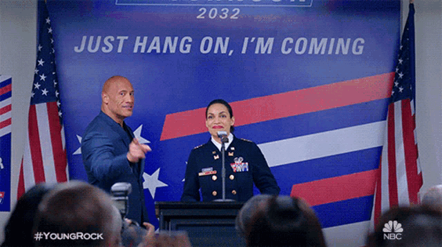 a man and a woman are giving a speech in front of a sign that says just hang on i 'm coming