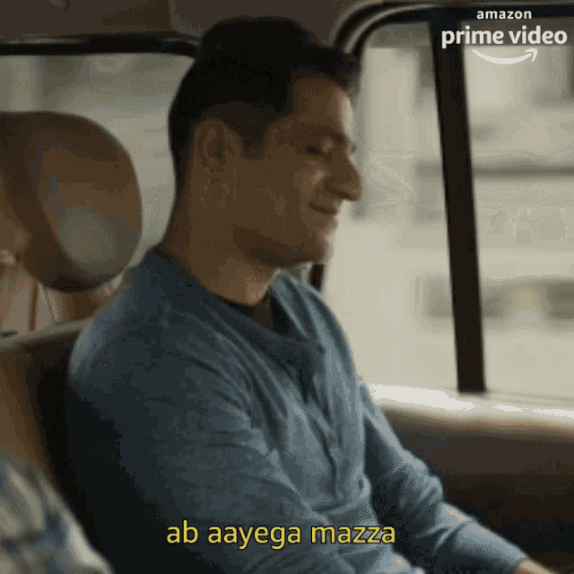 a man in a blue shirt is sitting in the back seat of a car and says ab aayega mazza .