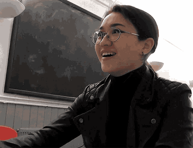 a woman wearing glasses and a black leather jacket smiles in front of a blackboard
